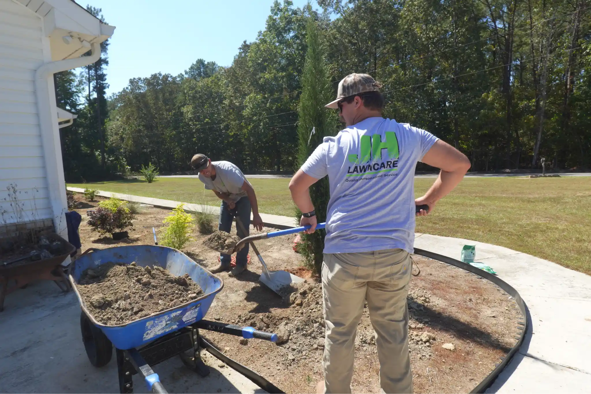 Two landscapers working on a yard near a house; one shovels dirt near a young cypress tree while a blue wheelbarrow filled with soil sits in the foreground